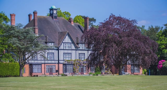 Das Tudor-Gebäude der Queenswood School steht umgeben von grünen Bäumen und einem Baum mit lila Blättern unter blauem Himmel.