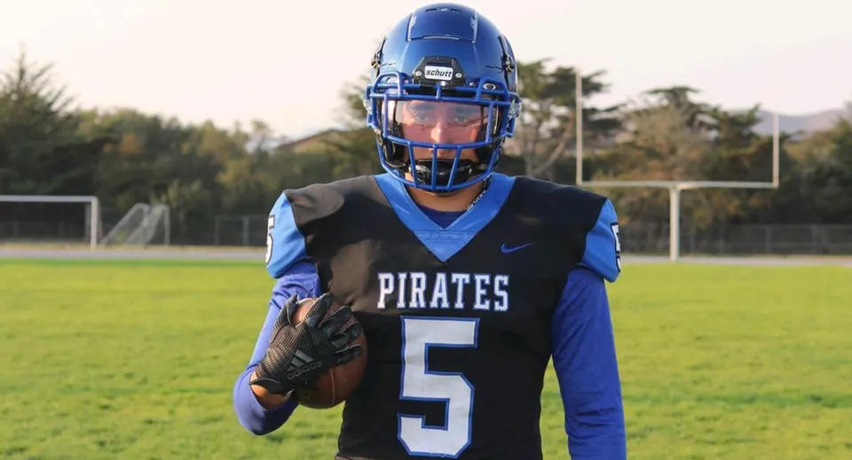 Uno studente del Mesa Unified School District indossa una maglia da football blu con il numero 5 e si trova su un campo verde davanti a un palo della porta.