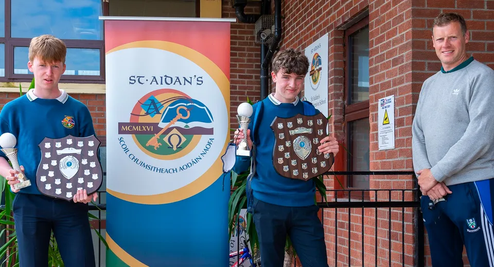Tre studenti della St. Aidan's Comprehensive School sono in piedi davanti a un edificio in mattoni con un grande cartello della scuola.