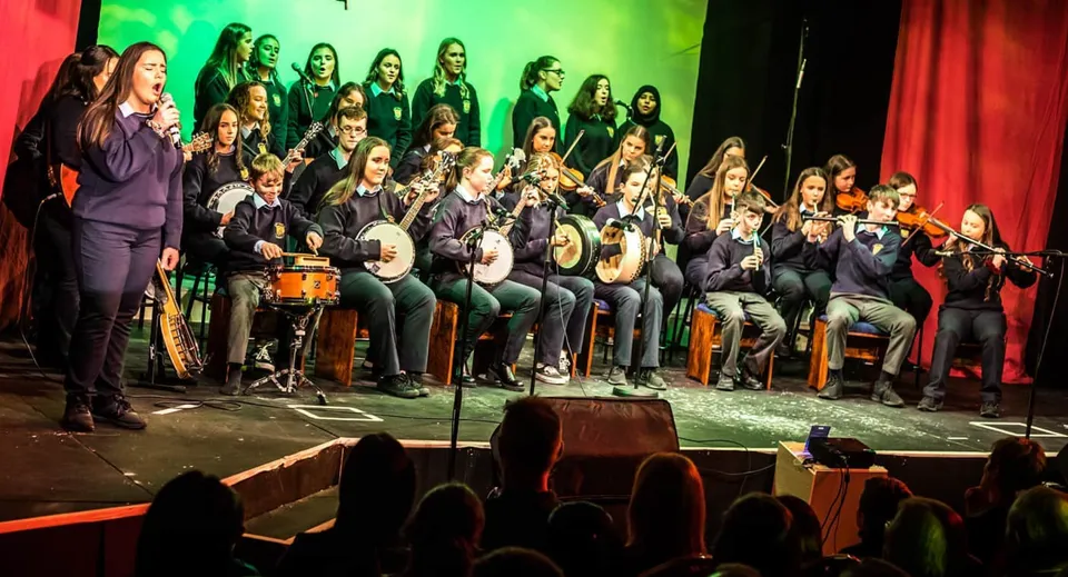 Il coro della Cnoc Mhuire Secondary School si esibisce su un palco con illuminazione colorata e sipario rosso.