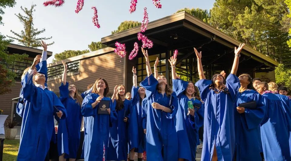 Un gruppo di neodiplomate e neodiplomati della Woodside Priory School indossa toghe blu davanti a una struttura in legno con fiori rosa pendenti.