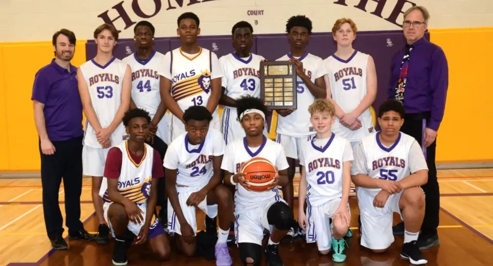 La squadra di basket della Centre Dufferin District High School posa insieme agli allenatori davanti alla parete con il nome e il logo della squadra, tenendo in mano un trofeo.