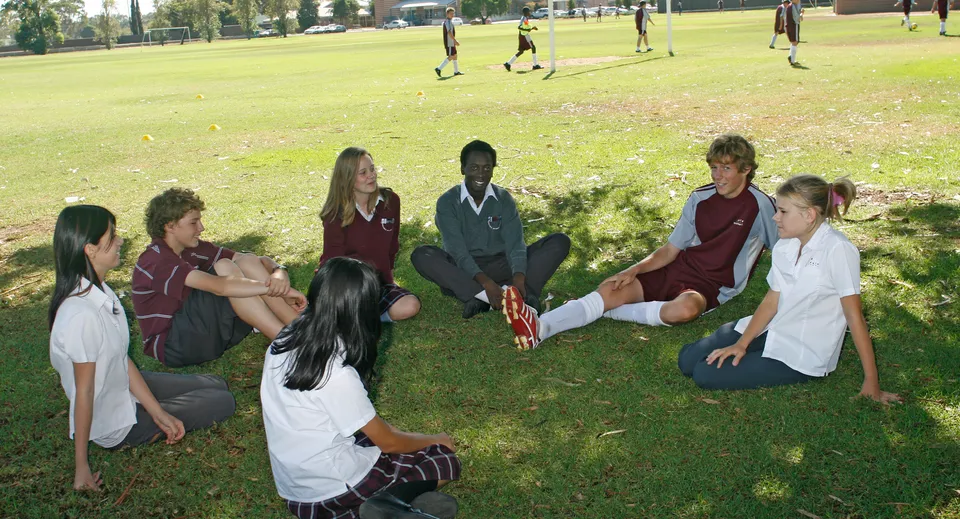 Studenti della Underdale High School siedono insieme su un prato in un'area simile a un parco con alberi sullo sfondo.