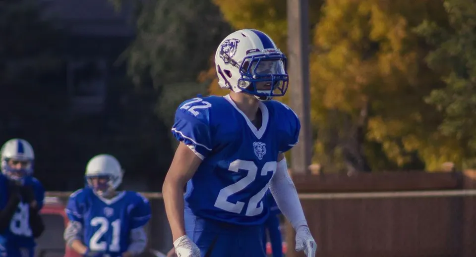 Un giocatore di football in maglia blu si trova di notte sul campo da gioco del River East Collegiate con altri giocatori sullo sfondo.