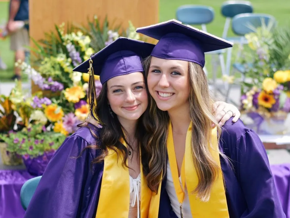Due diplomate della Warwick Valley High School posano sorridenti davanti a uno sfondo floreale colorato.
