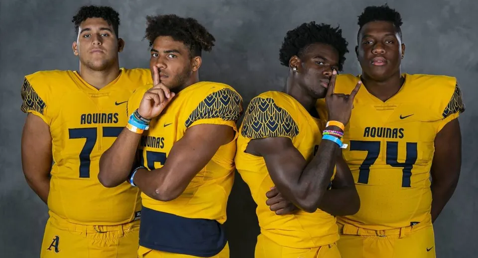 Quattro studenti della St. Thomas Aquinas High School indossano maglie gialle Aguias e posano insieme davanti a uno sfondo scuro.