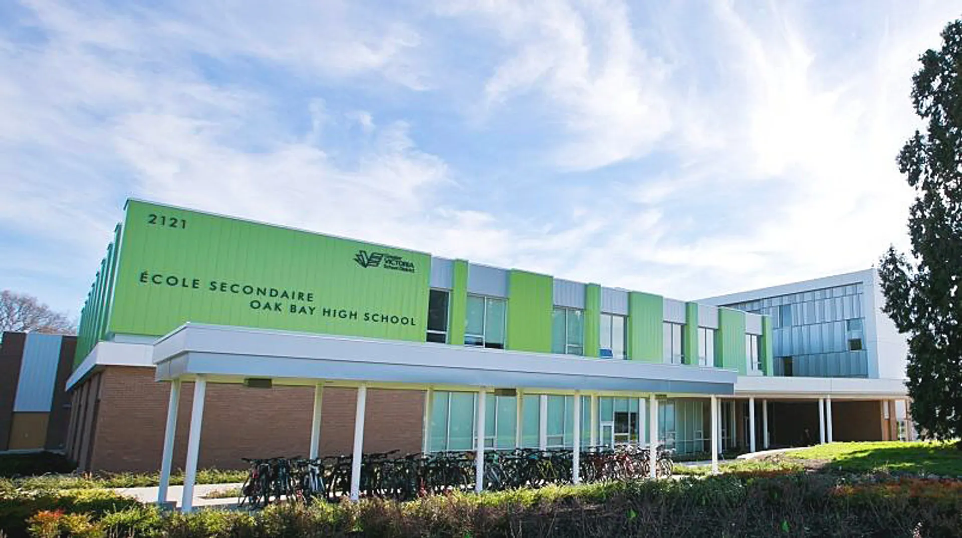 L'edificio scolastico moderno a più piani della Oak Bay Secondary High School, con facciata verde e grandi finestre, è circondato da alberi.