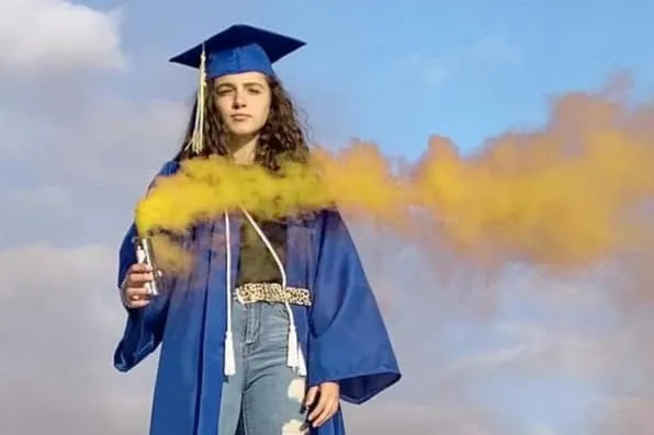 Joven con toga y birrete azules sosteniendo un bote con humo amarillo al aire libre