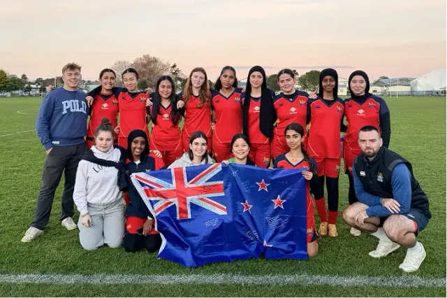 Squadra femminile in maglia rossa posa su un campo sportivo con la bandiera della Nuova Zelanda