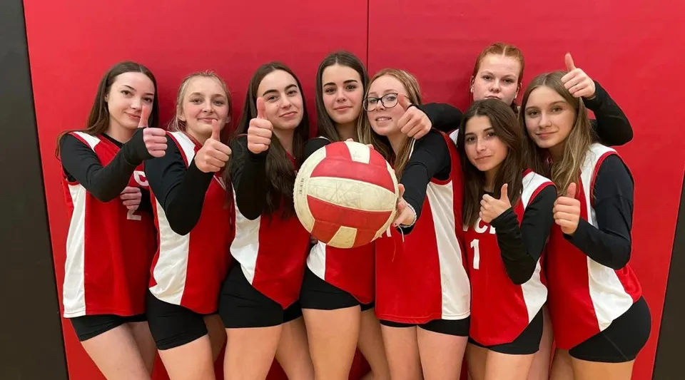 Un gruppo di giovani studentesse in maglia da pallavolo posa con un pallone davanti a uno sfondo rosso nel campus del Vankleek Hill Collegiate Institute.