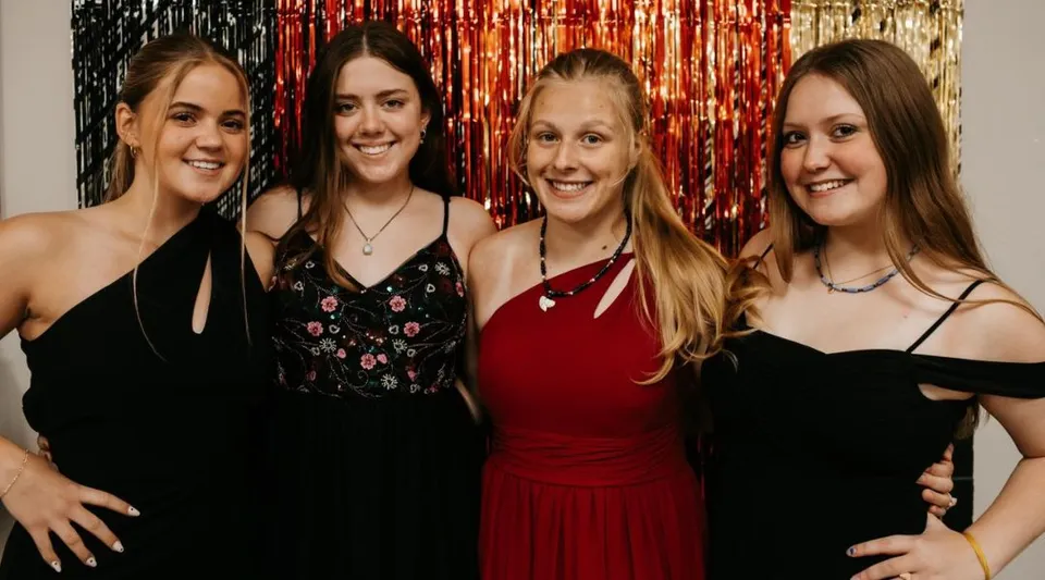 Quattro studentesse della Cambridge Christian School sorridono in vestiti eleganti davanti a uno sfondo luccicante.