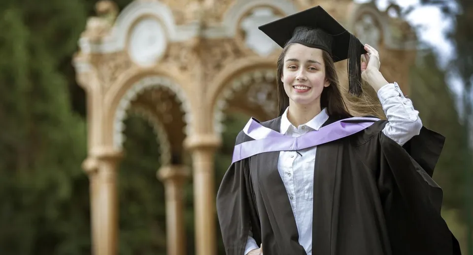 Una neodiplomata sorridente in toga e tocco è in piedi davanti a uno sfondo architettonico al Launceston College.