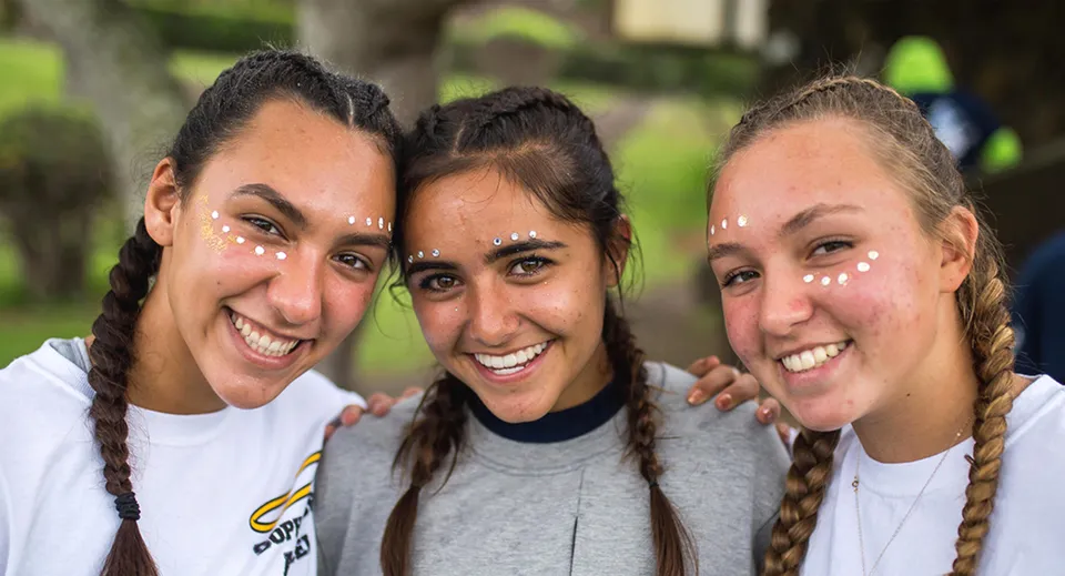 Tre studentesse della Hawaii Preparatory Academy sorridono insieme e posano all'aperto davanti ad alberi e verde.