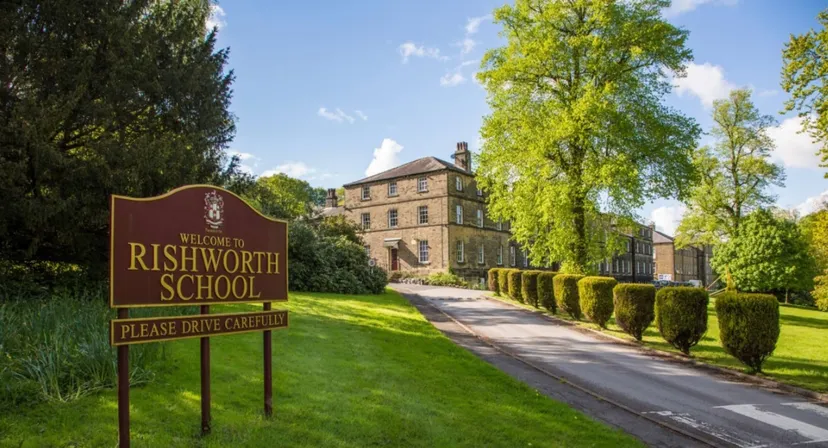 Il cartello d'ingresso della Rishworth School accanto a un viale che conduce a un edificio storico in pietra.
