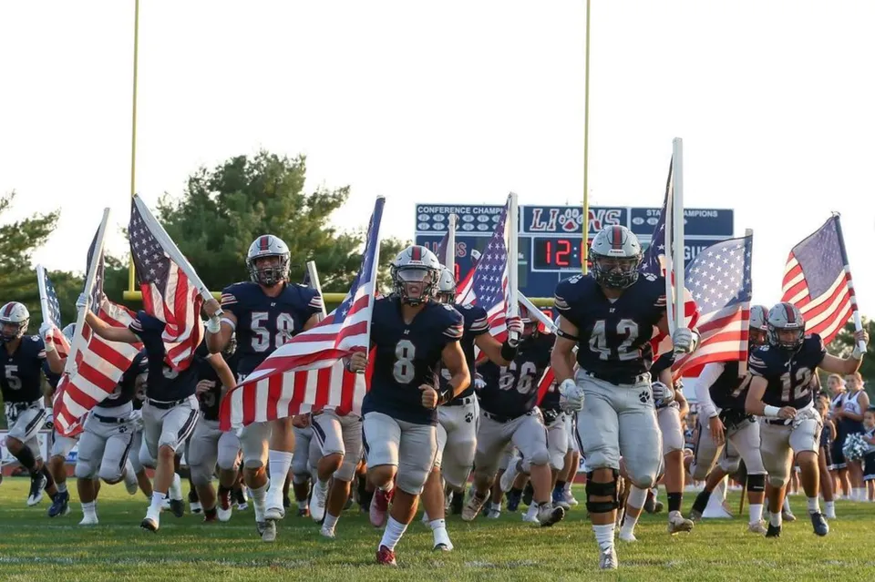 Un gruppo di giocatori di football corre in campo con bandiere americane alla Lacey Township High School.