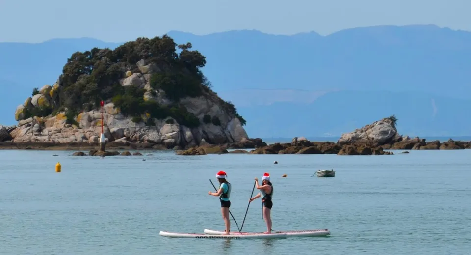 Due persone stanno in piedi su paddleboard in uno specchio d'acqua calmo davanti al paesaggio montuoso vicino alla Motueka High School.
