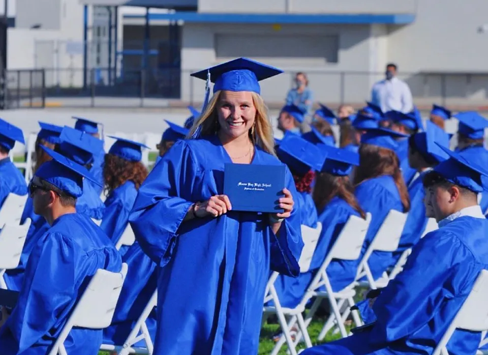 Una diplomata della Morro Bay High School con toga e tocco blu mostra il suo diploma accanto ai compagni.