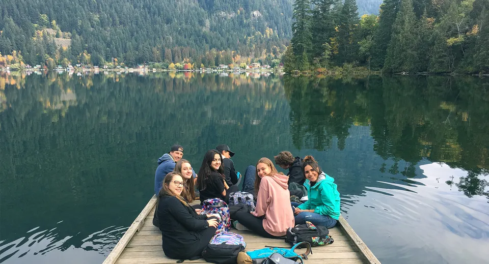 Un gruppo di studenti è seduto su un pontile di legno vicino a un lago con montagne boscose sullo sfondo presso il campus della Rick Hansen Secondary School.