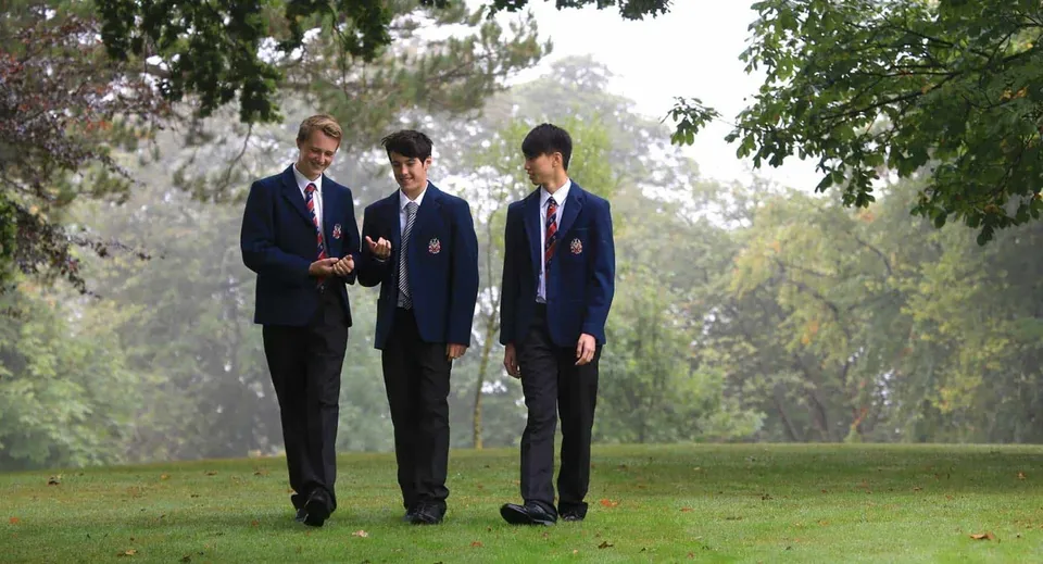 Tre studenti della Merchiston Castle School in uniforme scolastica camminano insieme su un prato all'aperto.