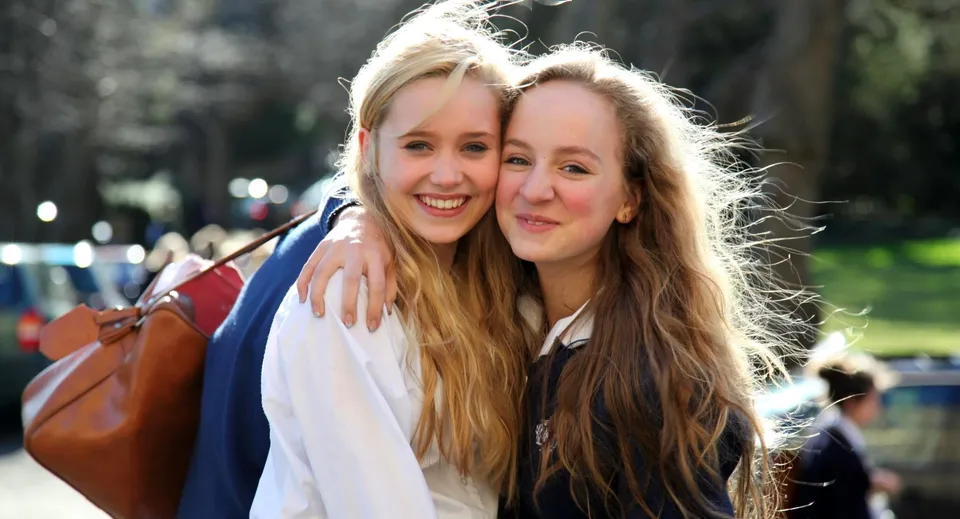 Due studentesse della Royal High School si abbracciano e sorridono in un'area esterna con alberi ed edifici.