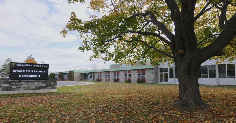 Un grande albero con foglie dai colori autunnali si trova davanti all'edificio scolastico della Rideau District High School con un avviso informativo per gli anni 7-8.