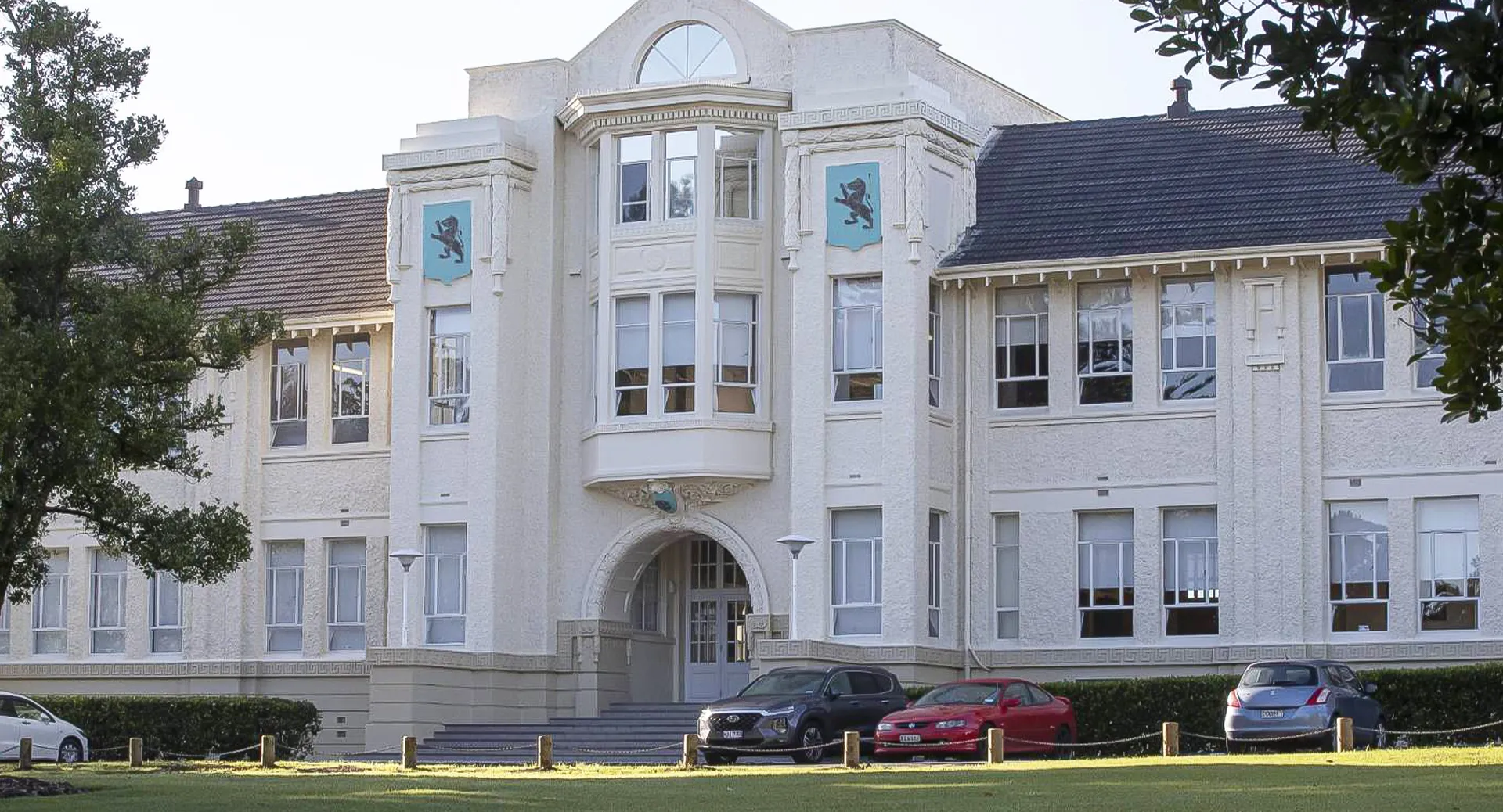 L'edificio bianco e decorato con finestre ad arco nel campus della Mount Albert Grammar School è circondato da alberi e auto parcheggiate.