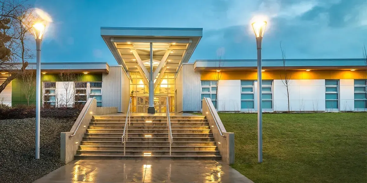 La entrada iluminada de la Georges Vanier Secondary School con una amplia escalera y dos farolas al atardecer.