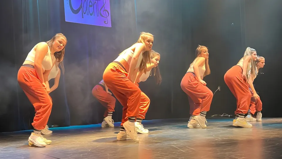 Quattro ballerini con outfit arancioni si esibiscono sul palco del Séminaire de Chicoutimi in uno spettacolo di danza con uno sfondo blu e viola.