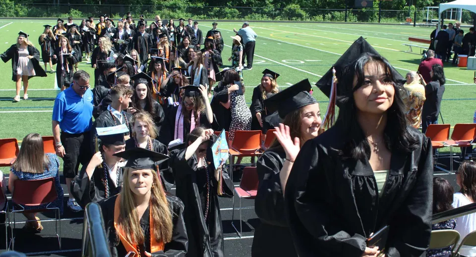 I diplomati del Marlborough College sono in piedi su un campo in toga nera e tocco, e posano in parte salutando.
