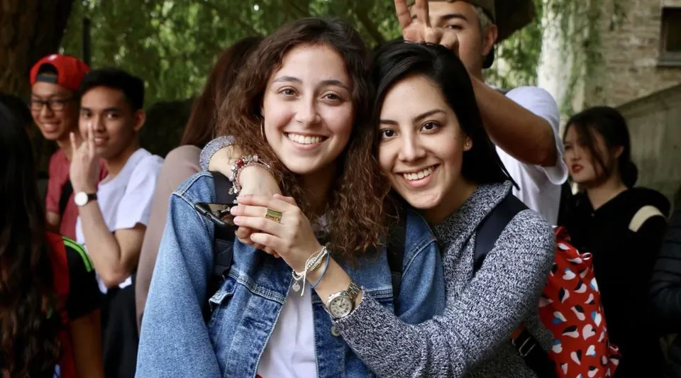 Due studentesse si abbracciano sorridendo in un gruppo nell'area verde all'aperto del St. Andrew’s College a Cambridge.