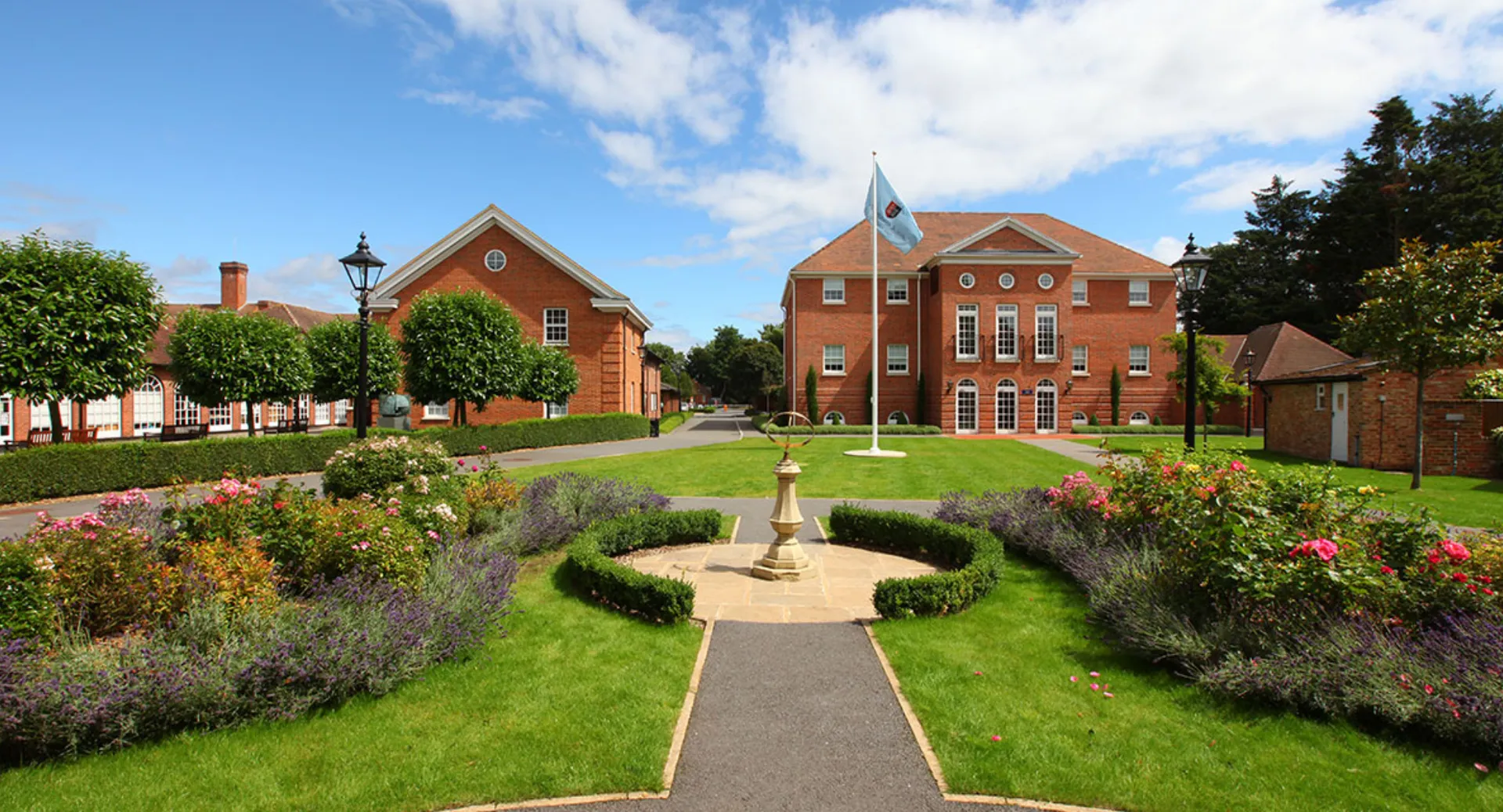 Il giardino ben curato con fontana conduce a un edificio in mattoni con torre dell'orologio nel campus della TASIS The American International School in England.
