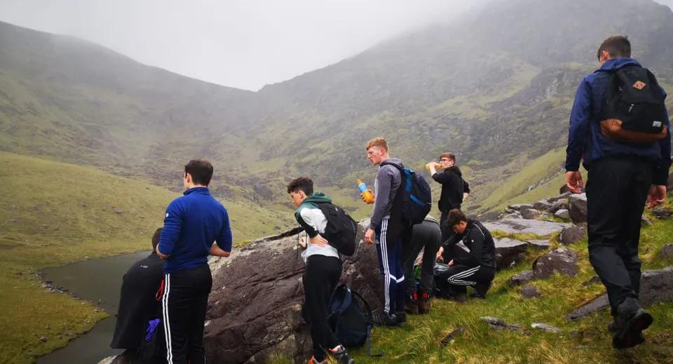 Studenti del St. Brendan's College sono in piedi in abbigliamento sportivo su una roccia con vista su un paesaggio montano nebbioso.