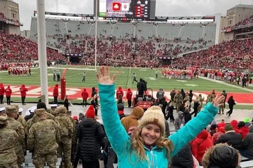 Joven con gorro y chaqueta turquesa levantando los brazos en la grada de un estadio