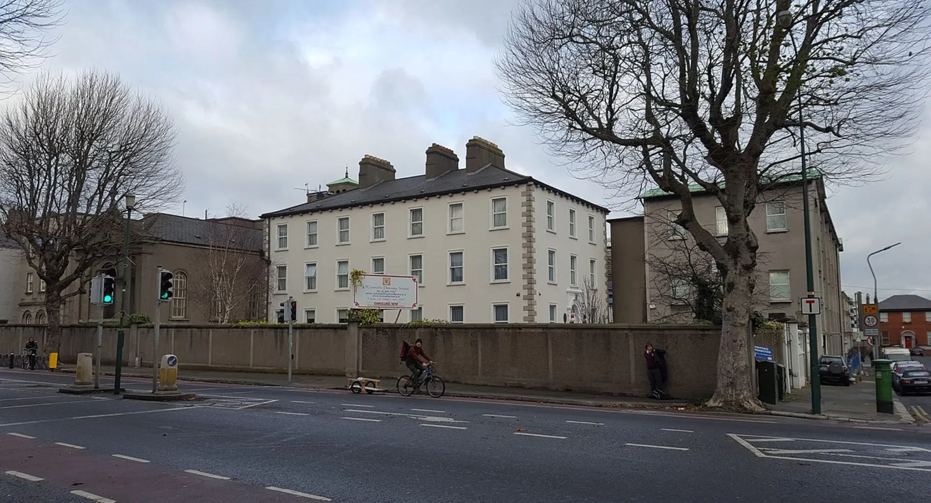 L'edificio a più piani del St. Mogues College è circondato da alberi spogli e in primo piano si trova una strada con un ciclista.
