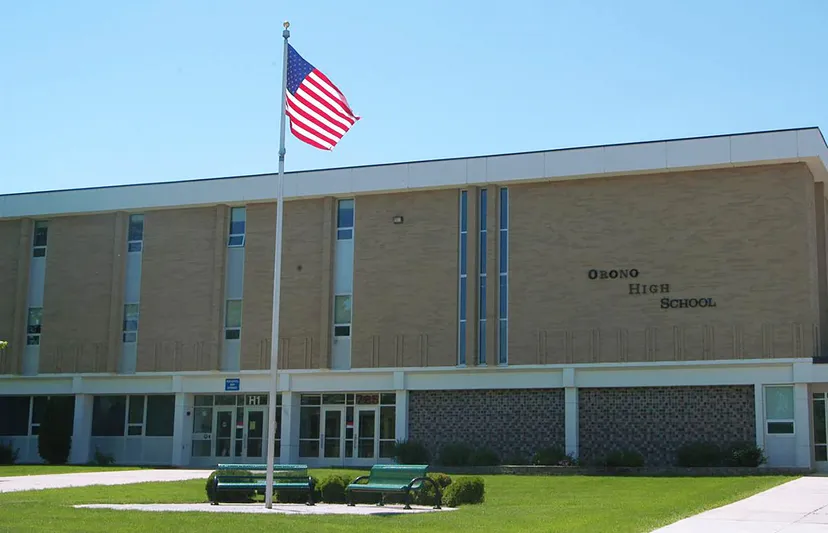 Il grande edificio in mattoni della Orono High School con una bandiera americana sul tetto e un prato con panchine davanti.
