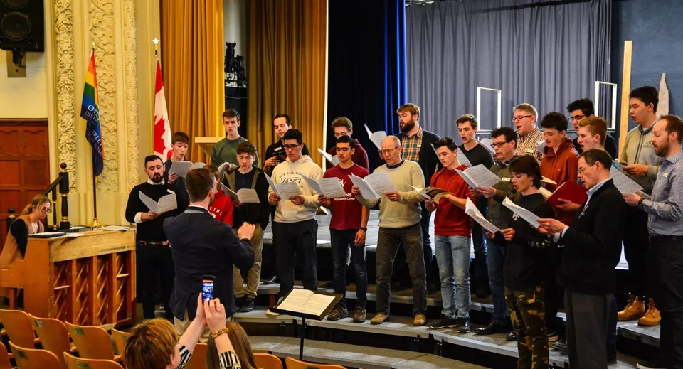 Un coro del Glebe Collegiate Institute si esibisce su un palco con elementi decorativi e bandiere sullo sfondo.