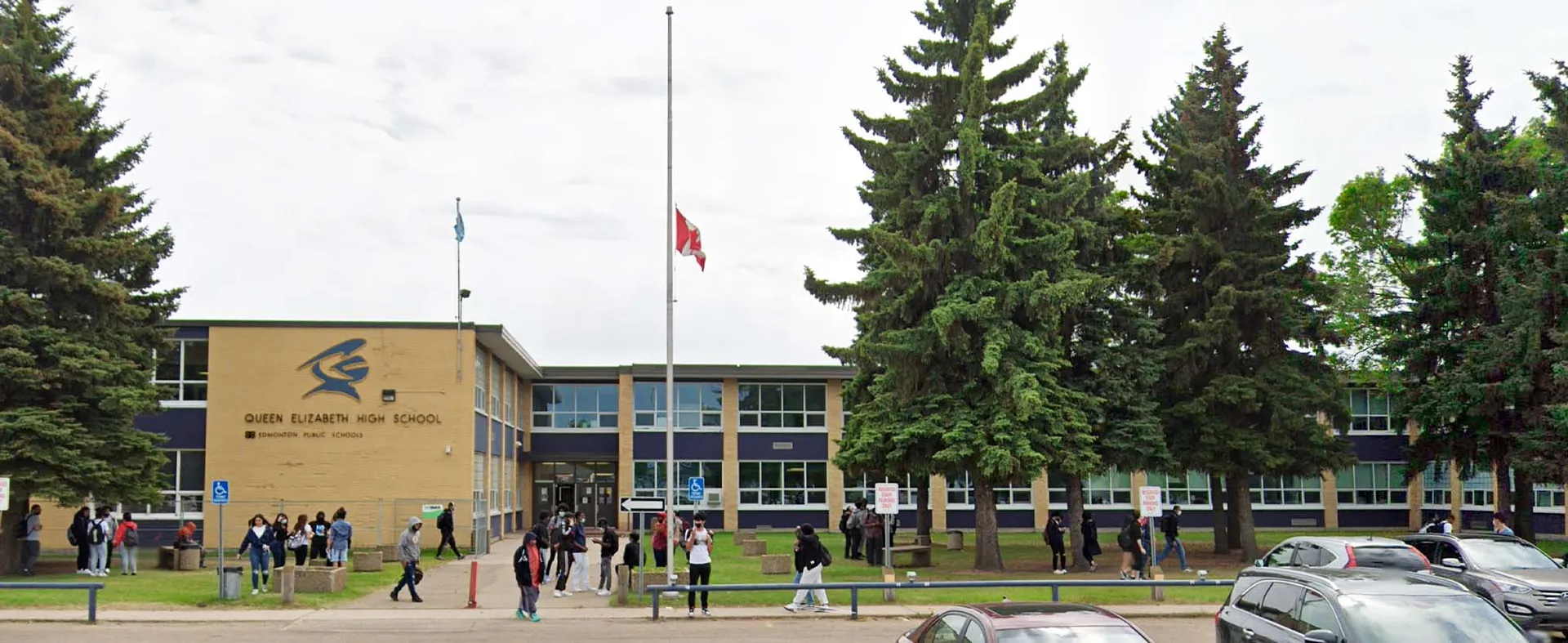 L'edificio scolastico della Queen Elizabeth High School circondato da alti abeti con un gruppo di studenti davanti e un pennone.