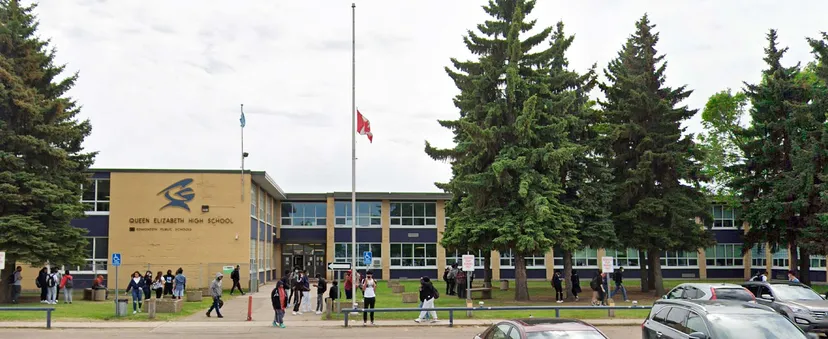 L'edificio scolastico della Queen Elizabeth High School circondato da alti abeti con un gruppo di studenti davanti e un pennone.
