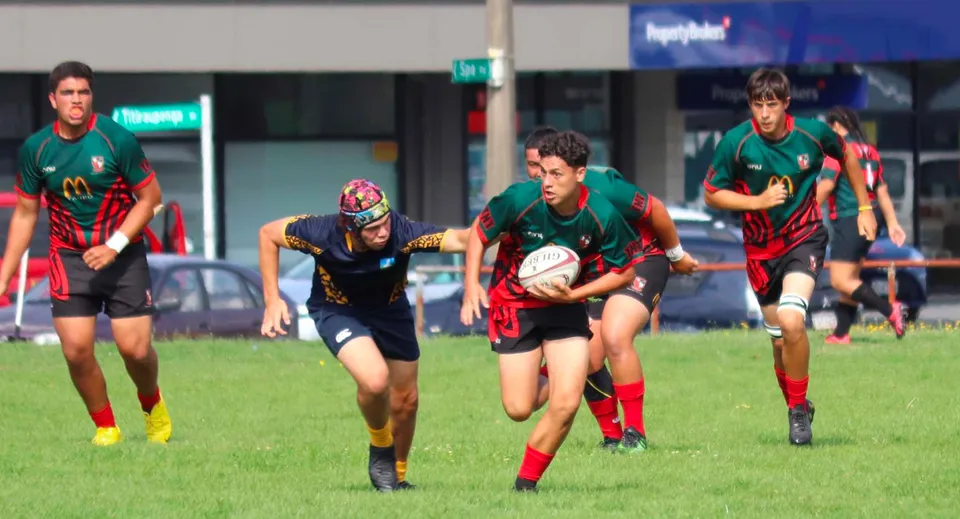 Studenti del Taupo-nui-a-Tia College giocano un'intensa partita di rugby su un campo con uno stadio sullo sfondo.