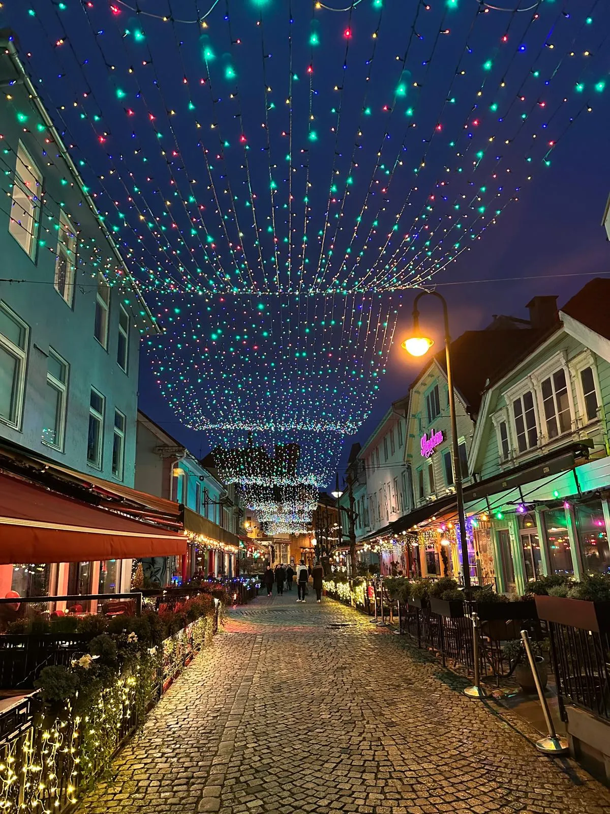 Via pedonale acciottolata addobbata con luci colorate tra caffè e locali all'imbrunire