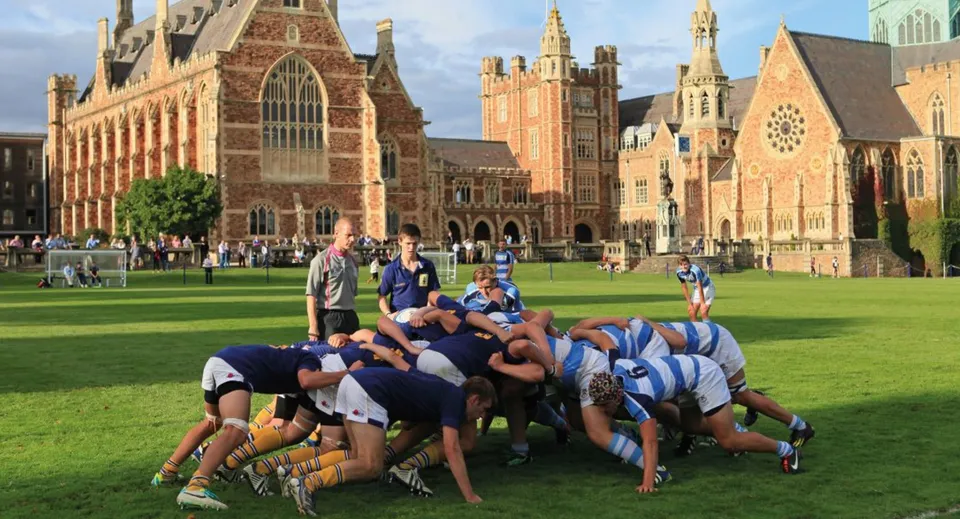 Un gruppo di studenti in abbigliamento sportivo forma un cerchio sul campo sportivo del Clifton College davanti a uno storico edificio in mattoni.