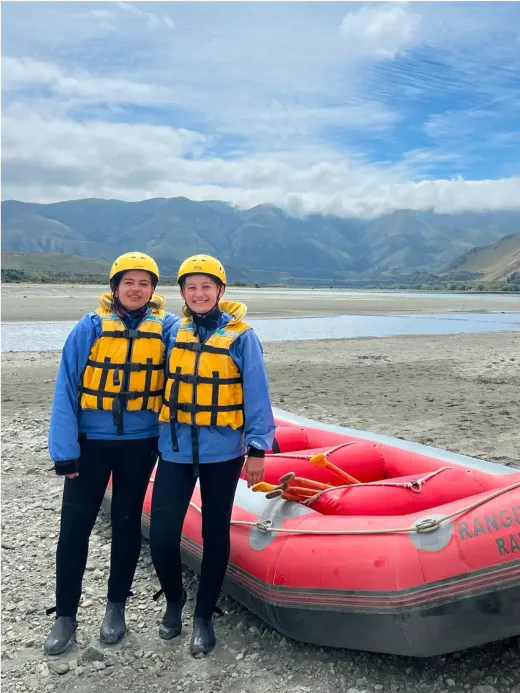 Zwei Mädchen mit Schwimmwesten und Helmen neben einem Raftingboot in Neuseeland