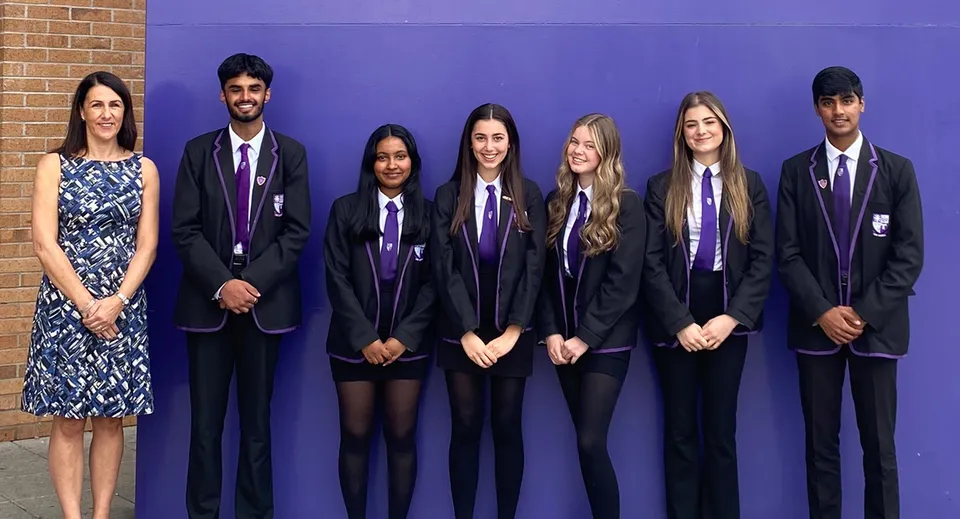 Un gruppo di studenti della Holy Rood High School in divisa scolastica formale è davanti a una parete viola con una donna in abito a fantasia.