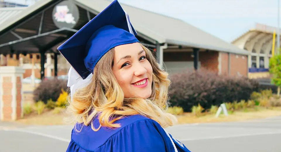 Una giovane diplomata della Queen Elizabeth High School è in piedi sorridente nella sua toga da diploma blu davanti a un edificio del campus.