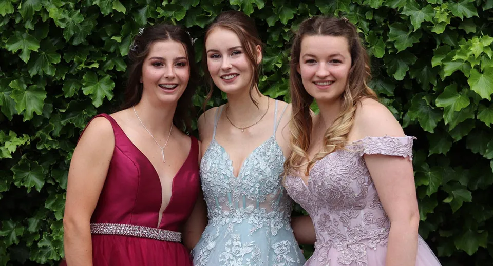 Tre studentesse della Queen Margaret's School for Girls si mettono in posa insieme davanti a un fogliame verde.