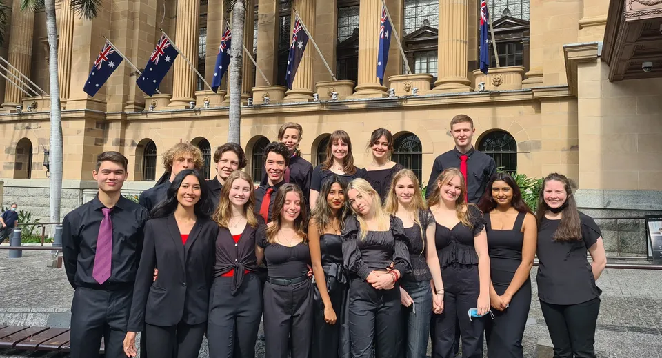 Un gruppo di studenti e studentesse della Indooroopilly State High School è in piedi in abito formale davanti a un grande edificio con colonne e bandiere australiane.
