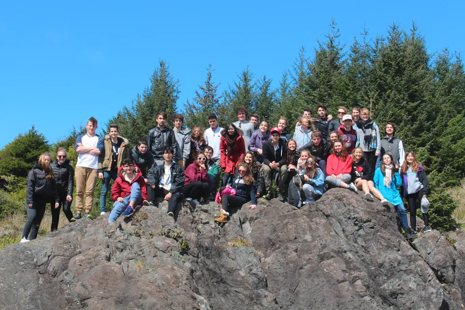 Un gruppo di studenti della Wellington Secondary School si trova su una roccia circondato da alberi sempreverdi sotto un cielo azzurro e limpido.