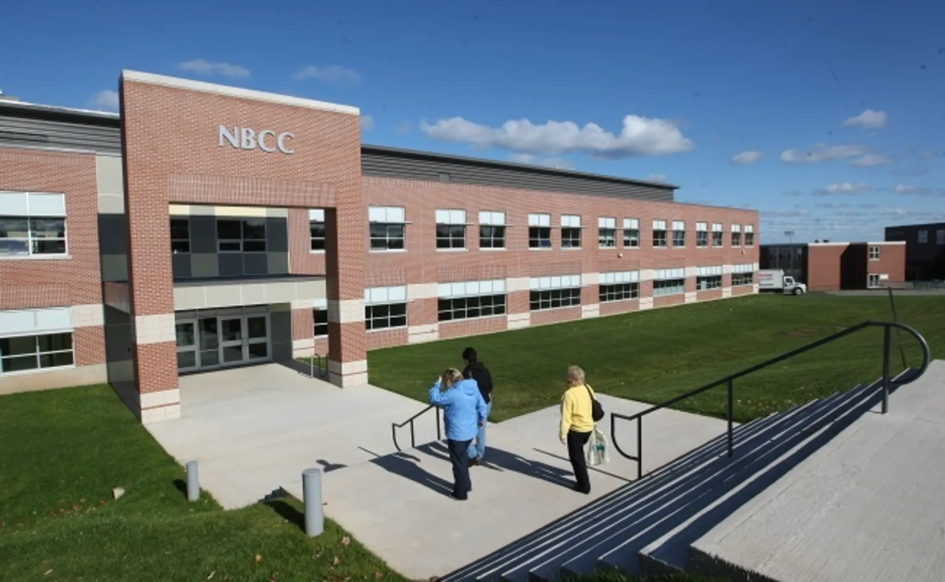 Due persone camminano su un sentiero davanti al grande edificio in mattoni del New Brunswick Community College sotto un cielo blu.