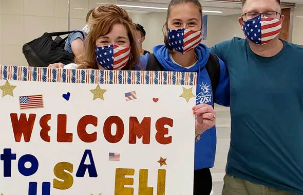Tres personas con mascarillas de la bandera de Estados Unidos sostienen un cartel de bienvenida