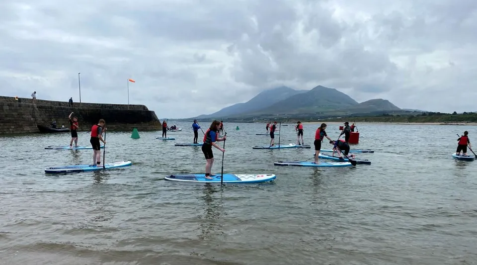 Studenti della Colaiste Mhuire (Tourmakeady) in piedi su paddle board in uno specchio d'acqua con montagne sullo sfondo.